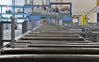 Blick entlang einer Rollenbahn in einer Industriehalle mit Regalen und Maschinen im Hintergrund