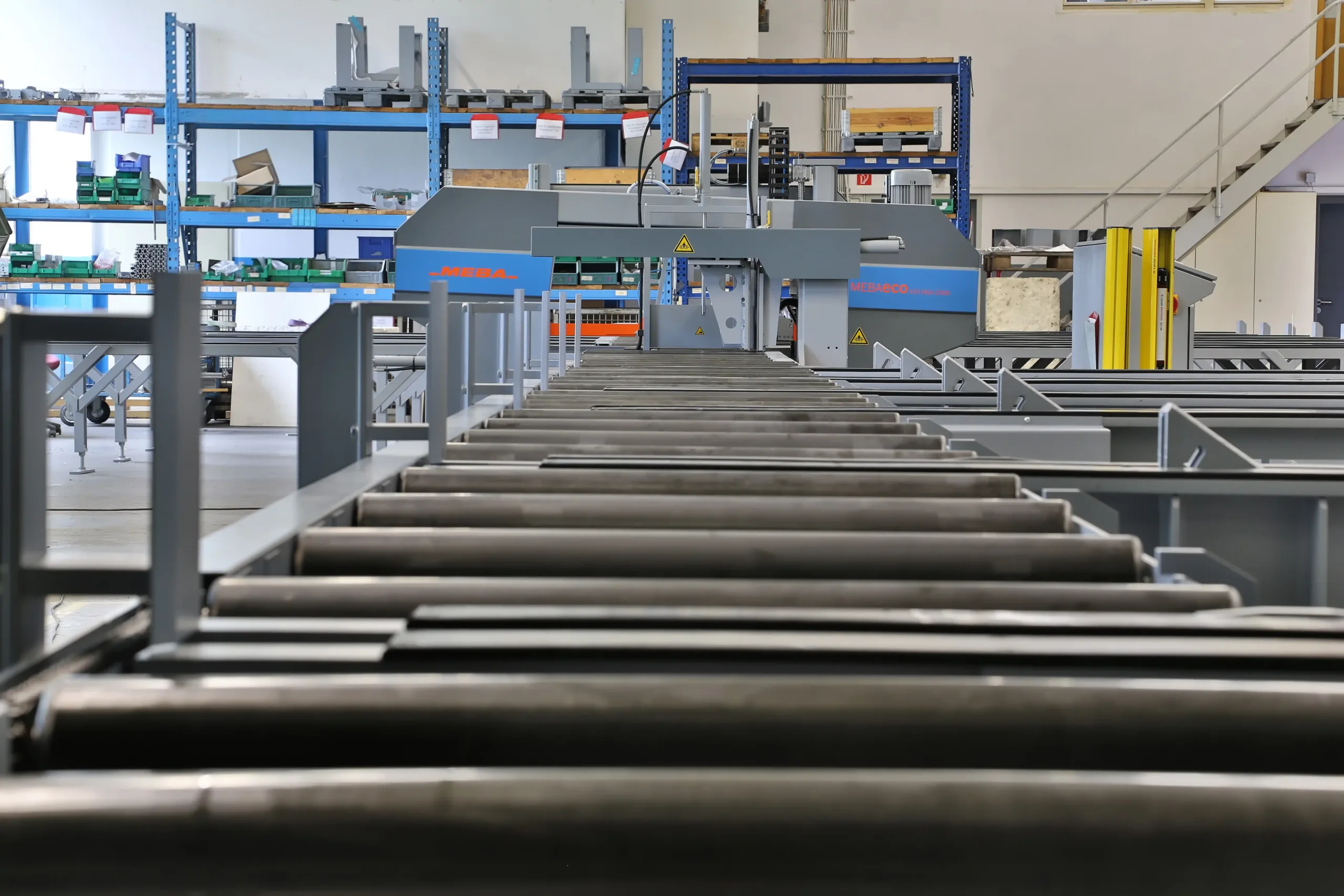 Blick entlang einer Rollenbahn in einer Industriehalle mit Regalen und Maschinen im Hintergrund